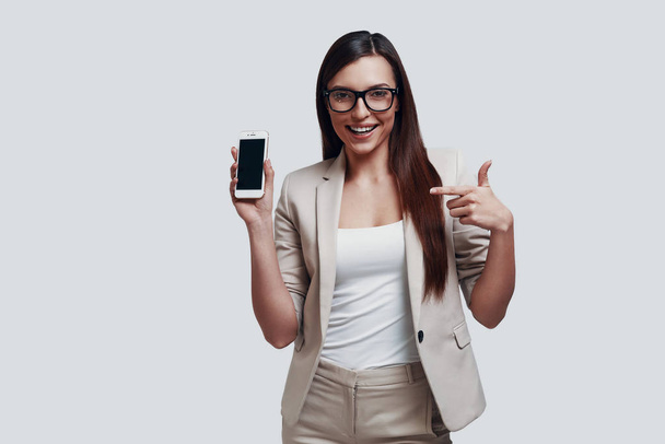 Nézz ide! Vonzó fiatal nő mutat másolás teret az okos telefon és mosolyogva állva ellen szürke háttér - Fotó, kép
