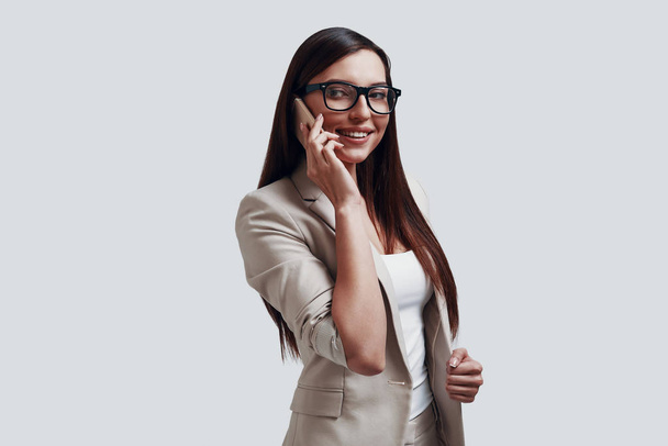 Good business talk. Attractive young woman talking on the phone and smiling while standing against grey background - Photo, Image