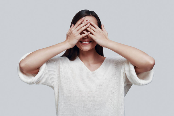 See nothing. Attractive young woman covering eyes with hands and smiling while standing against grey background - Photo, Image