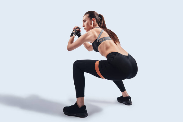Тяжело работать. Современная молодая женщина в спортивной одежде приседает с помощью бандажа сопротивления во время тренировок на сером фоне
 - Фото, изображение