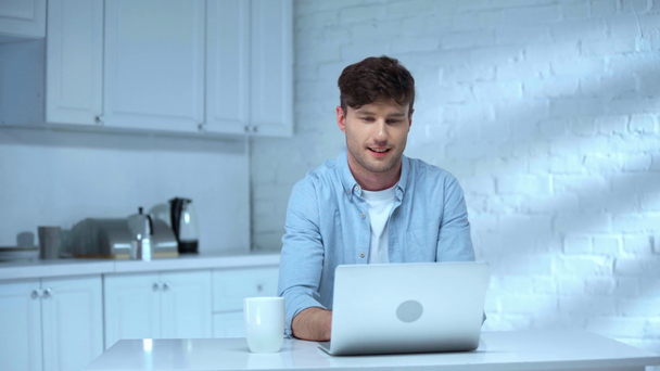 alegre freelancer usando laptop y bebiendo café con sonrisa en la cocina
 - Metraje, vídeo