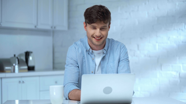 mosolygós szabadúszó használ laptopot asztalnál csésze kávét reggel a konyhában - Felvétel, videó