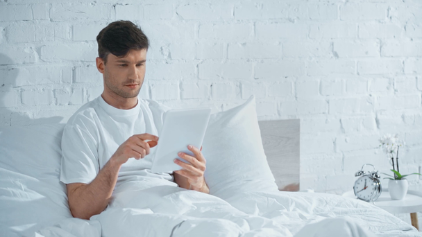 άντρας με λευκό μπλουζάκι ξαπλωμένο στο κρεβάτι, χρησιμοποιώντας ψηφιακό δισκίο και χαμογελώντας το πρωί - Πλάνα, βίντεο