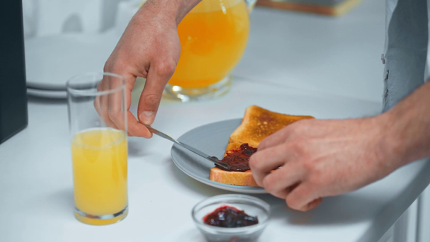 cropped view of man pouring orange juice in glass and applying jam on bread toasts - Materiaali, video