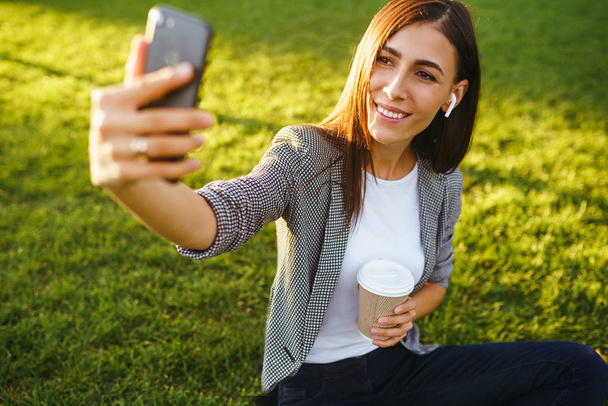 Time to take selfie! Image of beautiful stylish woman sitting on green grass with phone and coffee in the hands.She is talking on the phone through wireless headphones. Sunset light. Lifestyle concept - Valokuva, kuva