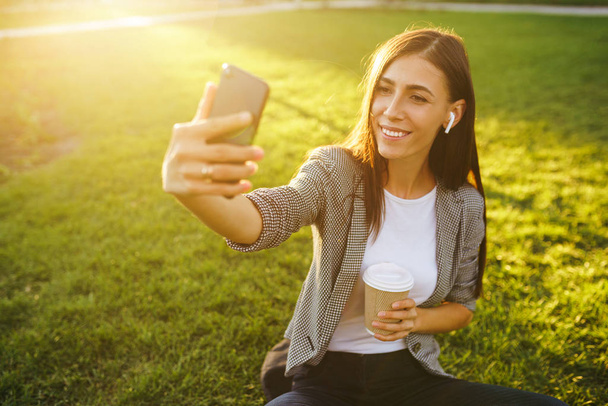 Time to take selfie! Image of beautiful stylish woman sitting on green grass with phone and coffee in the hands.She is talking on the phone through wireless headphones. Sunset light. Lifestyle concept - Zdjęcie, obraz