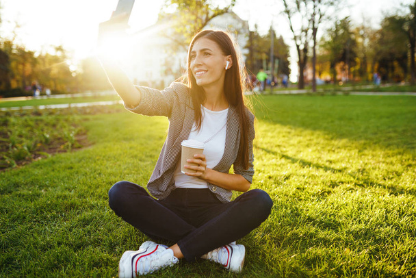 Zeit für ein Selfie! Bild von schönen stilvollen Frau sitzt auf grünem Gras mit Telefon und Kaffee in den Händen. Sie telefoniert über drahtlose Kopfhörer. Sonnenuntergang. Lifestylekonzept - Foto, Bild