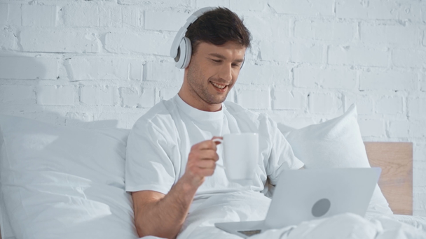 Freelancer w białym t-shirt picie kawy, słuchanie muzyki w słuchawkach i śmiech podczas pracy z laptopem w łóżku - Materiał filmowy, wideo