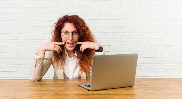 Jovem ruiva encaracolado mulher trabalhando com seu laptop sorri, apontando os dedos para a boca
. - Foto, Imagem
