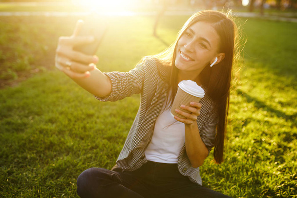 Time to take selfie! Image of beautiful stylish woman sitting on green grass with phone and coffee in the hands.She is talking on the phone through wireless headphones. Sunset light. Lifestyle concept - Photo, image