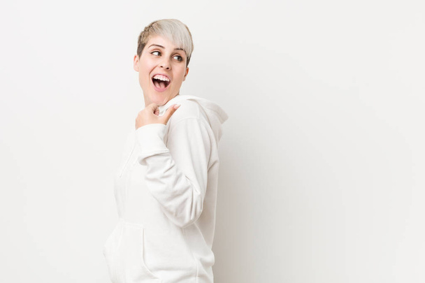 Jeune femme courbée portant un sweat à capuche blanc pointe avec le pouce loin, riant et insouciant
. - Photo, image