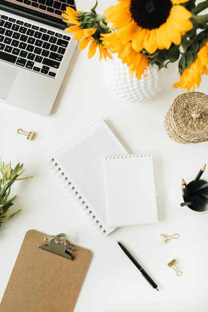 moderner Schreibtisch mit Laptop, Notizbuch, gelbem Sonnenblumenstrauß auf weißem Hintergrund. flache Lage, Draufsicht Freelancer / Blogger Sommer florales Arbeitskonzept. - Foto, Bild