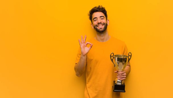 Young man holding a trophy cheerful and confident doing ok gesture - 写真・画像