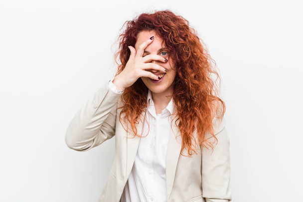 Young natural redhead business woman isolated against white background blink at the camera through fingers, embarrassed covering face. - Photo, Image
