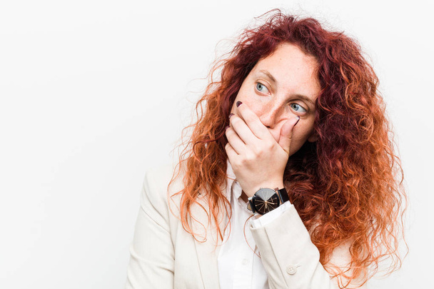 Jeune femme d'affaires rousse naturelle isolée sur fond blanc réfléchi regardant vers un espace de copie couvrant la bouche avec la main
. - Photo, image