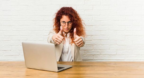 Νεαρή κοκκινομάλλα σγουρή γυναίκα που δουλεύει με το laptop της με σηκωμένους τους αντίχειρες, ζητωκραυγάζει για κάτι, υποστήριξη και σεβασμό. - Φωτογραφία, εικόνα