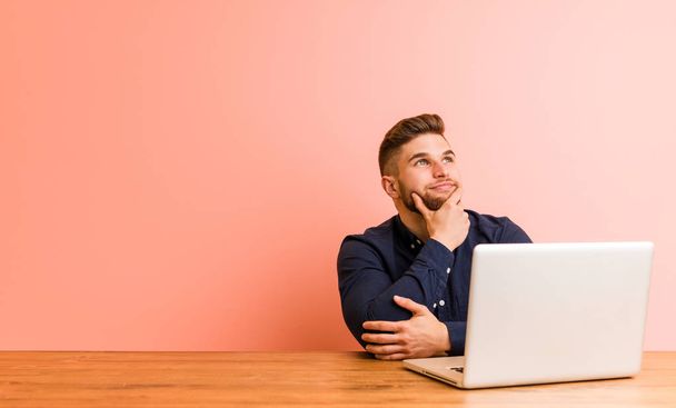 Jeune homme travaillant avec son ordinateur portable regardant latéralement avec une expression douteuse et sceptique
. - Photo, image
