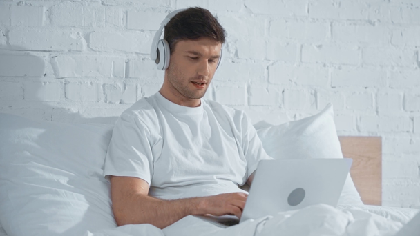 ανεξάρτητος σε λευκό μπλουζάκι ακούγοντας μουσική στα ακουστικά και δουλεύοντας με το laptop, ενώ ξαπλωμένο στο κρεβάτι - Πλάνα, βίντεο