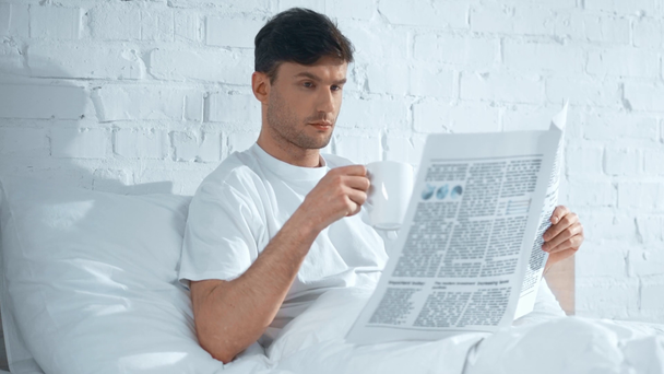 koncentrovaný muž s bílým tričkem, popíjení kávy a čtení novin v posteli - Záběry, video