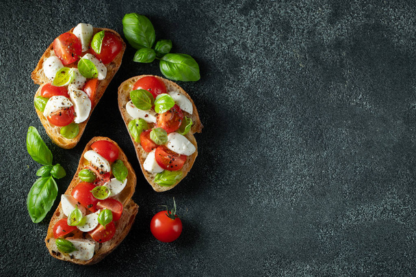 暗い背景にトマト、モッツァレラチーズ、バジルのブルスケッタ。伝統的なイタリアの前菜やスナック、antipasto。コピースペース付きの上面図。フラットレイ - 写真・画像