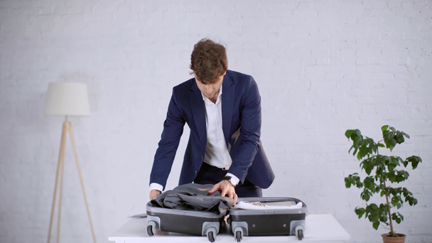 jóképű üzletember csomagolás ruhákat és záró bőrönde az asztalra otthon - Felvétel, videó
