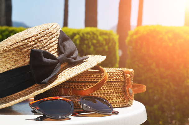 Круглий солом'яний мішок, солом'яний капелюх та чорні сонцезахисні окуляри з зеленими кущами та блакитним небом на фоні сонячного світла
 - Фото, зображення