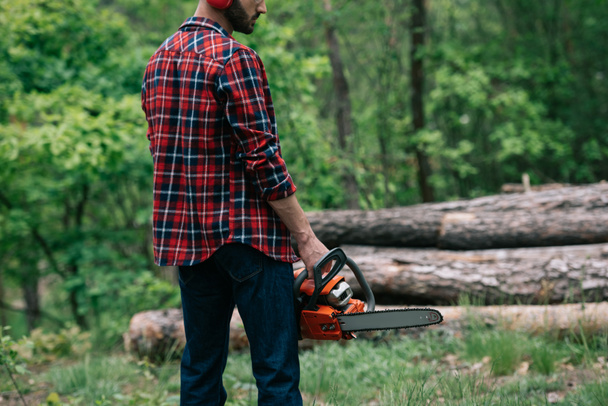 oříznutý pohled na dřevorubec v Kostkové košili držící motorovou pilu v lese - Fotografie, Obrázek