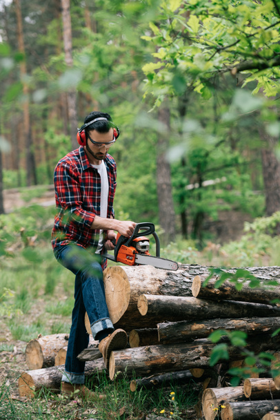 lumberman in earmuffs and protective glasses cutting trunks with chainsaw in forest - Photo, Image