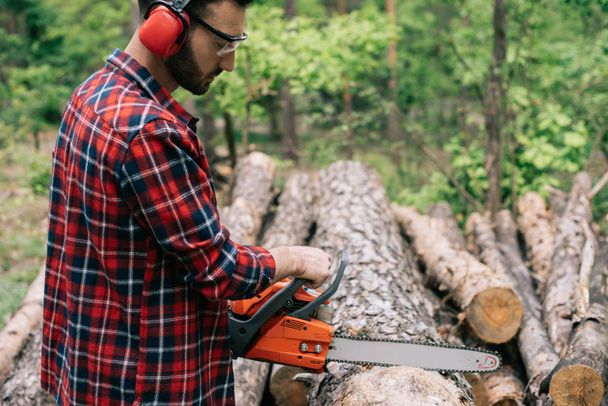 concentrated lumberer in earmuffs and protective glasses cutting wood with chainsaw in forest - Фото, изображение