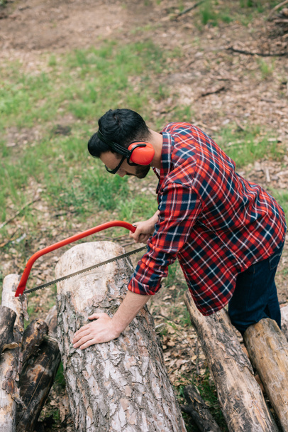 lumberjack in checkered shirt and noise-canceling headphones cutting log with handsaw in forest - Photo, Image