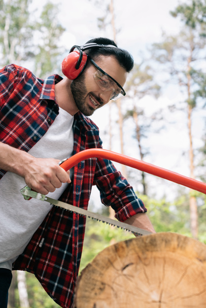 smiling lumberjack in earmuffs cutting tree trunk with bowsaw in forest - Photo, Image