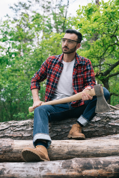 thoughtful lumberman holding ax while sitting on tree trunks in forest and looking away - Photo, Image