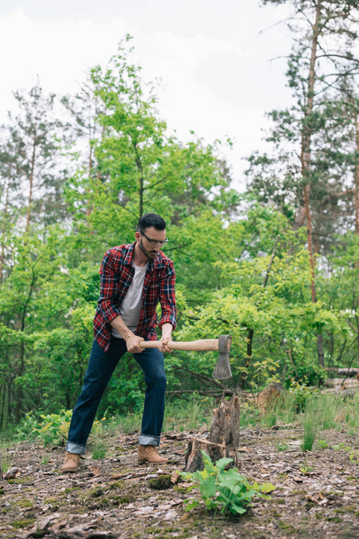 lumberjack in checkered shirt and denim jeans cutting wood with ax in forest - Valokuva, kuva
