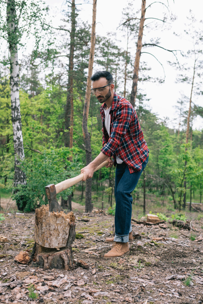 handsome lumberjack in plaid shirt and denim jeans cutting wood with ax in forest - Zdjęcie, obraz