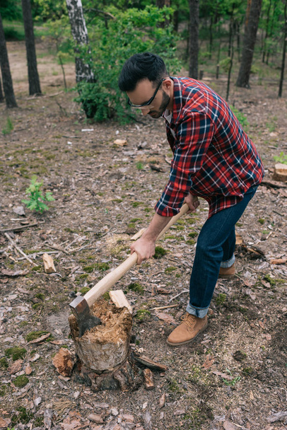 lumberjack in plaid shirt and denim jeans cutting wood with ax in forest - Foto, Imagem