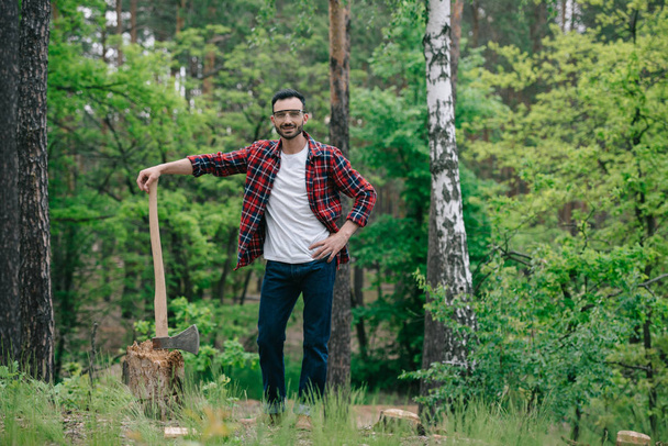 smiling lumberjack in plaid shirt and denim jeans holding ax while standing with hand on hip and looking at camera - Fotografie, Obrázek