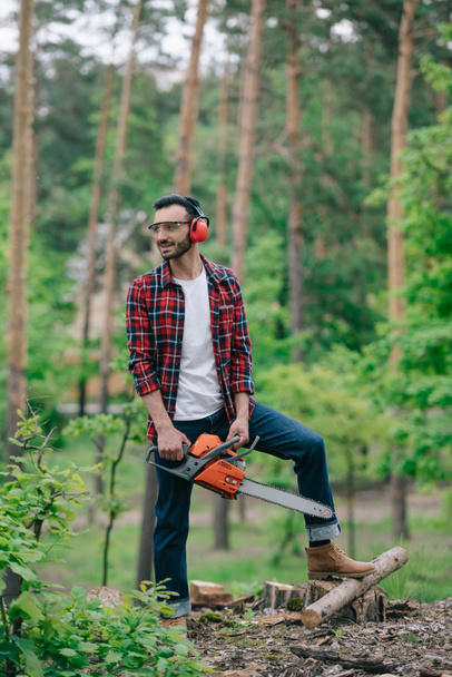 boscaiolo sorridente in camicia a quadri e jeans in denim in piedi con motosega nel bosco e distogliendo lo sguardo
 - Foto, immagini