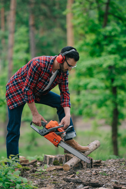 lumberer in noise-canceling headphones cutting wood with chainsaw in forest - Photo, Image