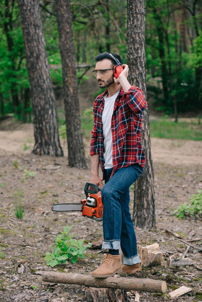 thoughtful lumberjack holding chainsaw, touching noise-canceling headphones and looking away - Photo, Image