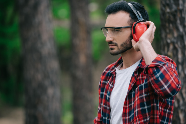 pensive lumberjack touching noise-canceling headphones and looking away in forest - Photo, Image