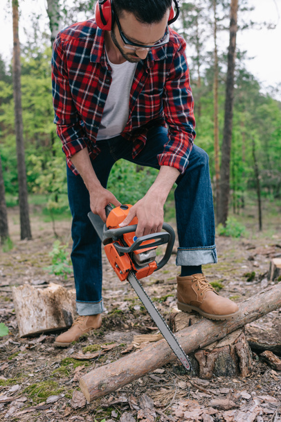 lumberer in geruite overhemd en denim jeans snijden hout met kettingzaag in bos - Foto, afbeelding
