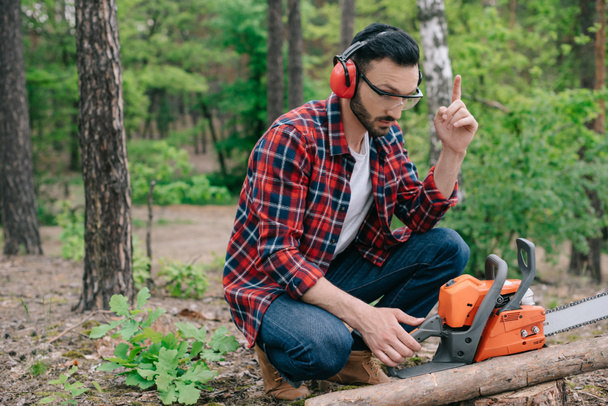 lumberjack in noise-canceling headphones sitting in forest near chainsaw and showing idea gesture - Photo, Image