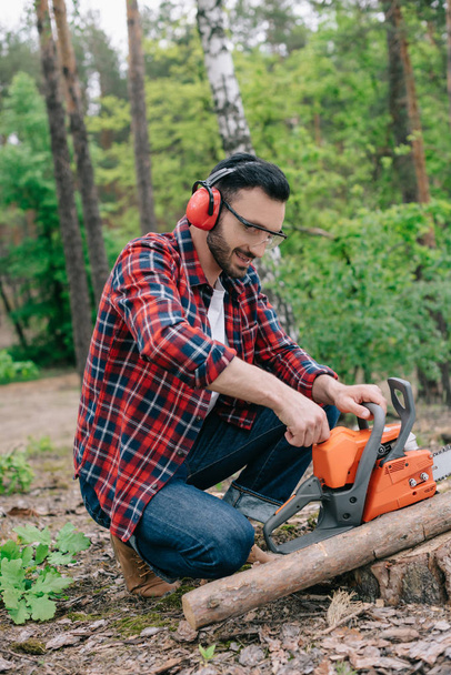 lumberjack in noise-canceling headphones adjusting chainsaw in forest - Foto, afbeelding