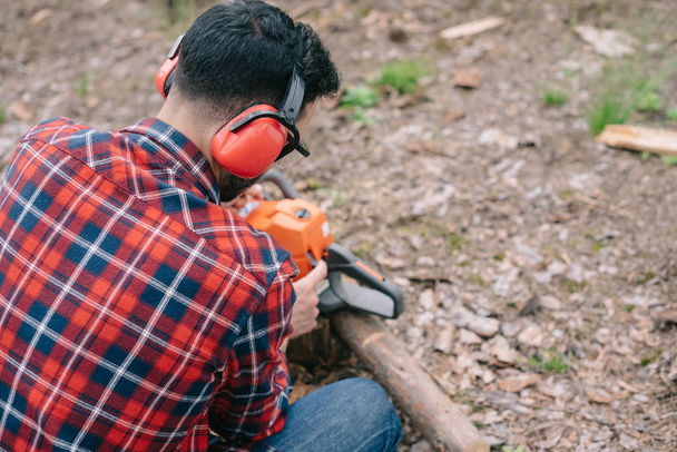 back view of lumberjack in hearing protectors repairing chainsaw in forest - Photo, Image