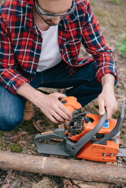 partial view of lumberman in plaid shirt adjusting chainsaw in forest - Foto, Imagen