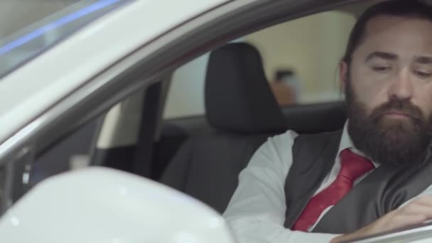 Portrait attractive confident bearded man sitting in the vehicle and inspects newly purchased auto from the car dealership. Car showroom. Advertising concept. - Video
