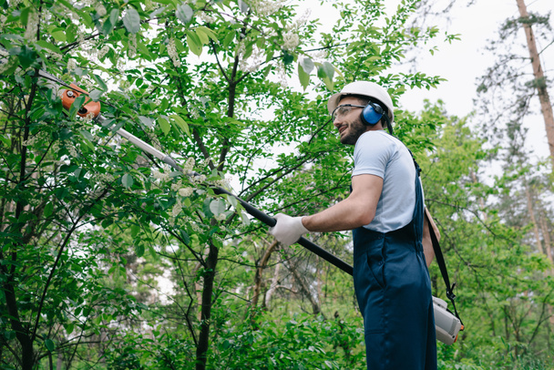 улыбающийся садовник в комбинезоне и слуховые протекторы, обрезающие деревья с помощью телескопической шестовой пилы в парке
 - Фото, изображение
