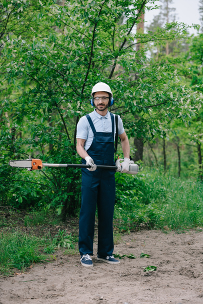 hermoso jardinero en overoles, casco y orejeras sosteniendo la sierra telescópica y mirando a la cámara
 - Foto, imagen