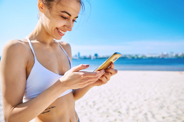 Closeup portrait of beautiful sexy happy woman on the sand city beach with sea background, looking at phone and smiling. Summertime concept. - Zdjęcie, obraz