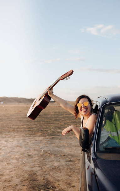 Mujer viajero joven divirtiéndose con la guitarra en el coche jeep 4x4 haciendo unas vacaciones de angostura al atardecer
 - Foto, imagen